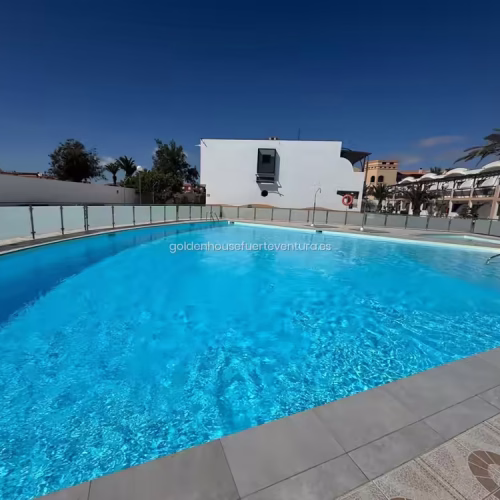 Piscina comunitaria vallada con agua azul y zona de ducha exterior bajo el sol.