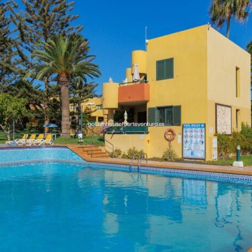 Piscina junto a edificio de dos plantas color amarillo con palmeras de fondo.