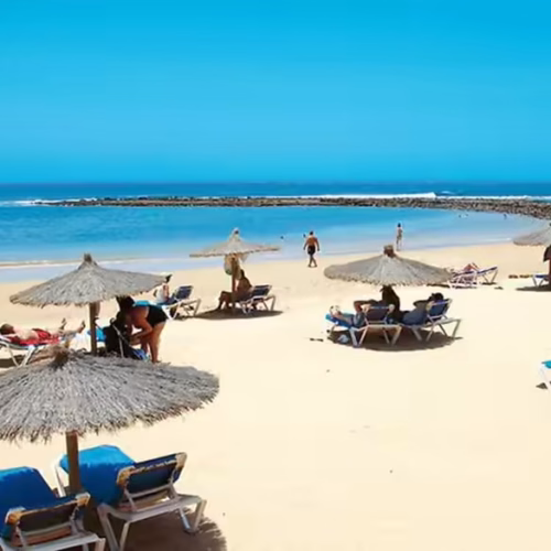 Playa de arena fina con sombrillas de brezo, hamacas azules y espigón de rocas al fondo.