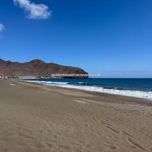 Vista amplia de la playa de arena oscura y aguas tranquilas bajo cielo azul.