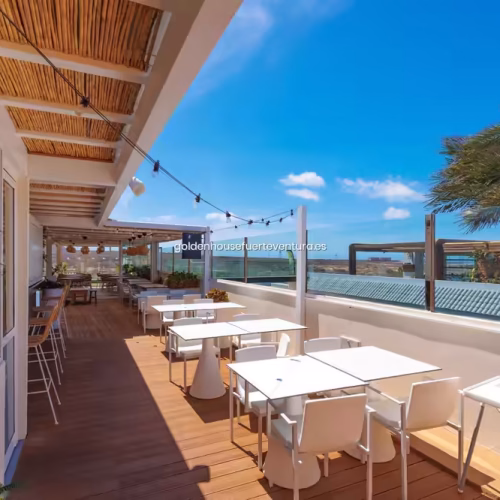 Zona de comedor exterior cubierta con mesas blancas, sillas modernas y techo de madera con luces decorativas.