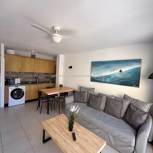 Vista del salón con sofá gris, mesa de comedor de madera y cocina al fondo.