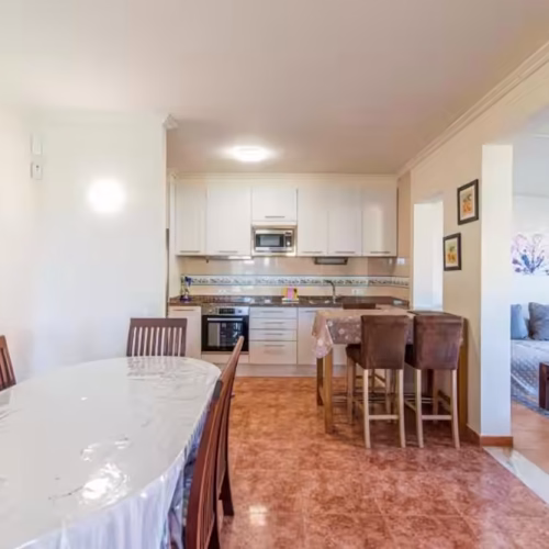 Mesa de comedor de madera para seis personas integrada con cocina blanca y zona de estar.