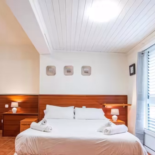 Cama de matrimonio con cabecero de madera, mesitas de noche y ventanal luminoso en Fuerteventura.