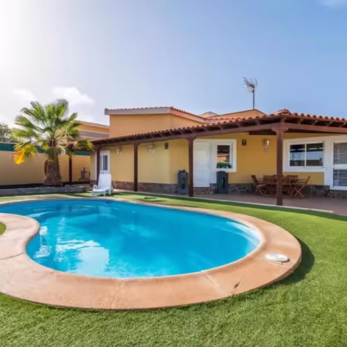 Fachada de la villa de color amarillo con piscina privada en forma de riñón y zona de porche.