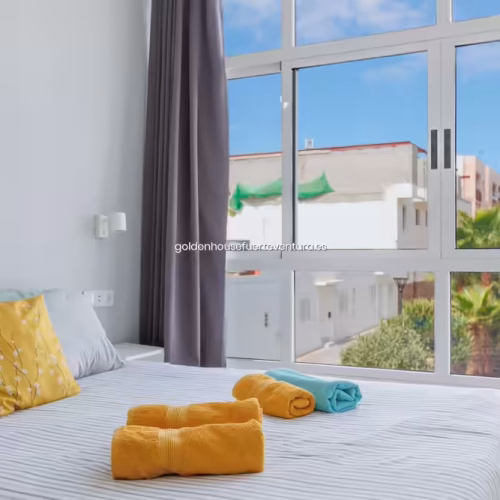 Perspectiva desde la cama hacia el ventanal mostrando edificios cercanos y palmeras