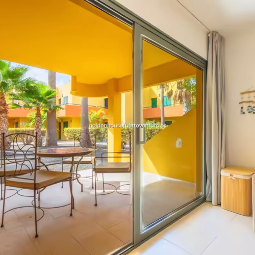 Puerta corredera acristalada con vistas a un jardín con palmeras y edificios amarillos.